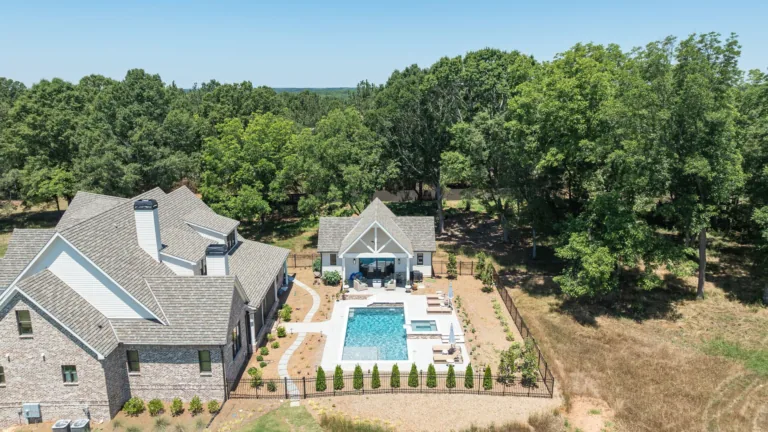 Aerial view of a stone house with a backyard pool, patio, and surrounding trees in a wooded setting