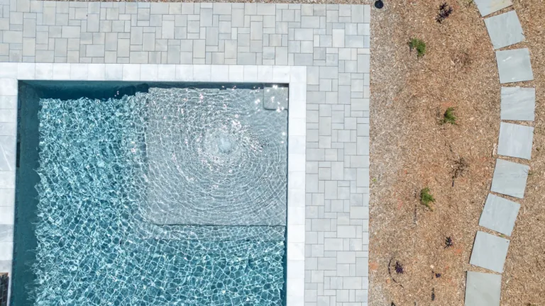 Aerial view of a pool with light stone deck beside a curved gravel path and sparse landscaping.