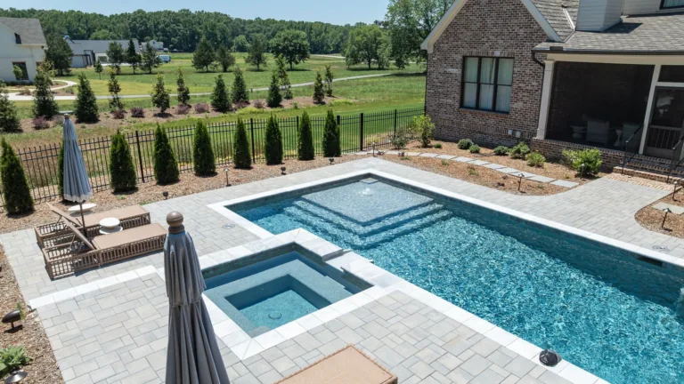 Backyard pool with raised spa, stone patio, lounge chairs, and landscaped lawn beside a house