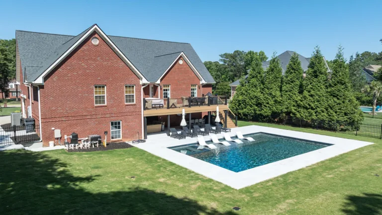 Brick house with a backyard pool, patio chairs, and green lawn under a clear blue sky