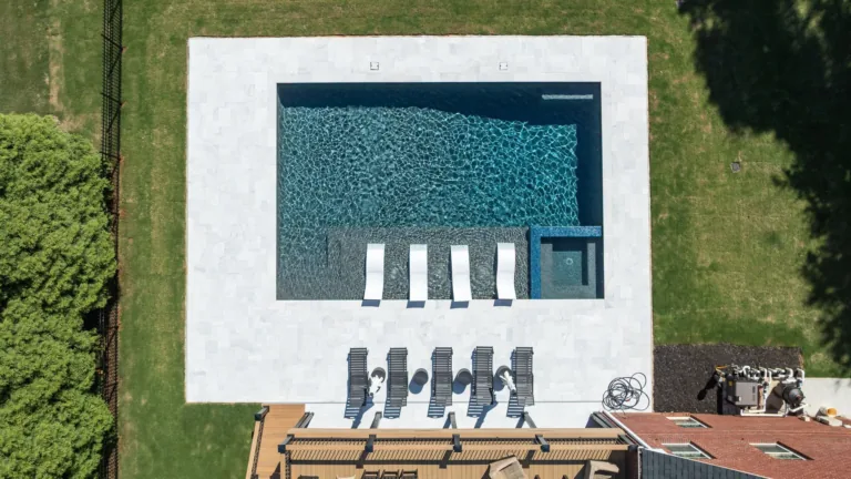 Aerial view of a rectangular swimming pool with white lounge chairs and a patio beside green grass.