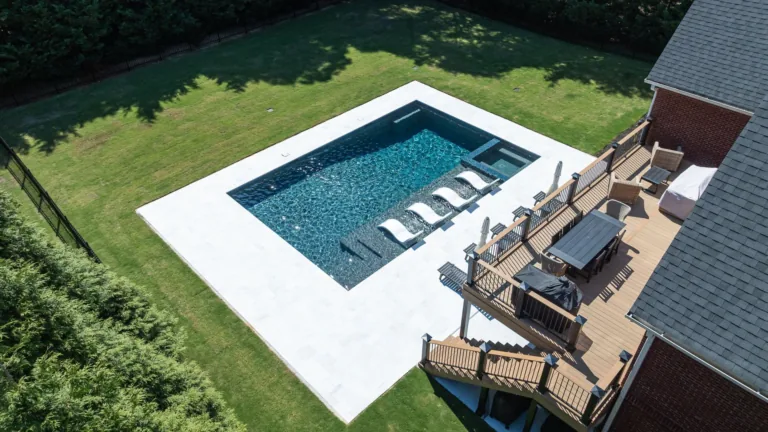 Backyard pool with lounge chairs beside a wooden deck and a brick house, surrounded by green lawn.