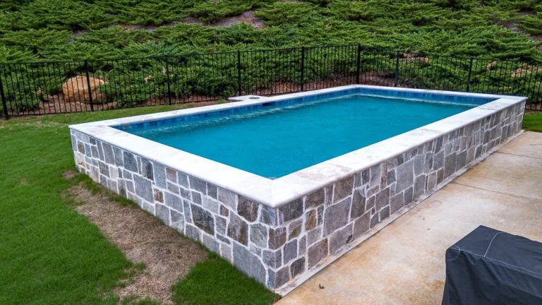 Rectangular pool with stone walls, surrounded by grass, concrete, and black fence.