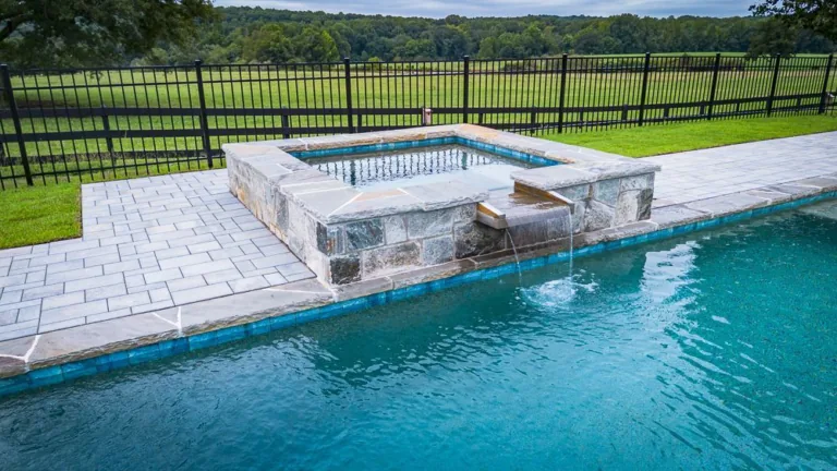 Pool with stone hot tub and waterfall, black fence, and green field.