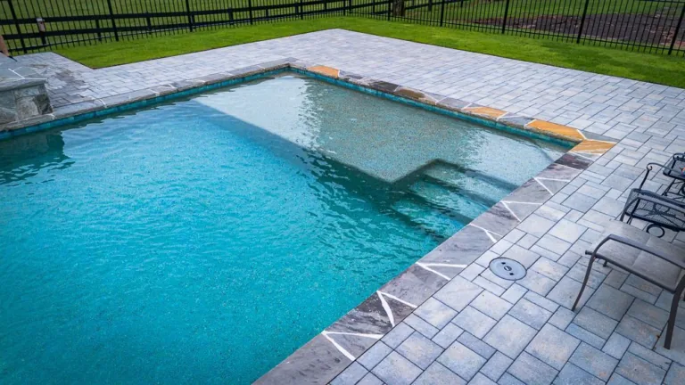 A rectangular swimming pool with gray brick patio, grass, and a fence in the background.