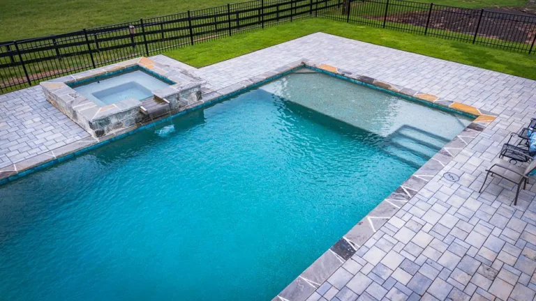Outdoor pool with attached hot tub, surrounded by stone patio. Blue water, green grass and black fence.