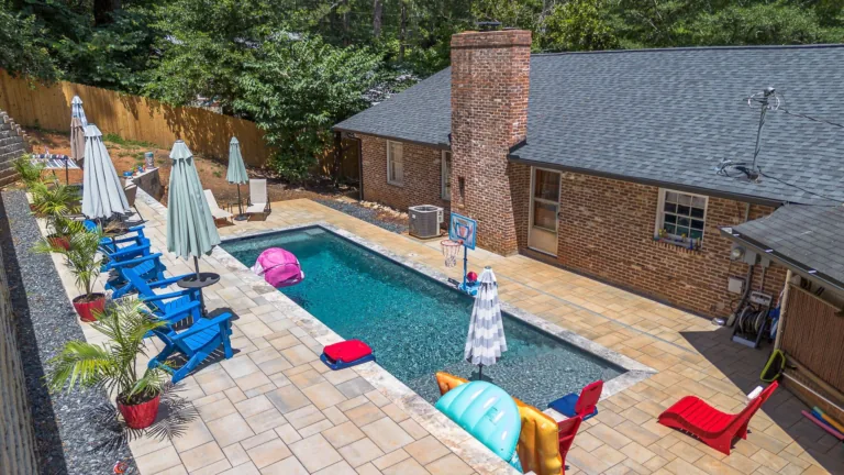Backyard pool with blue chairs, umbrellas, and inflatables. Brick building with a chimney.