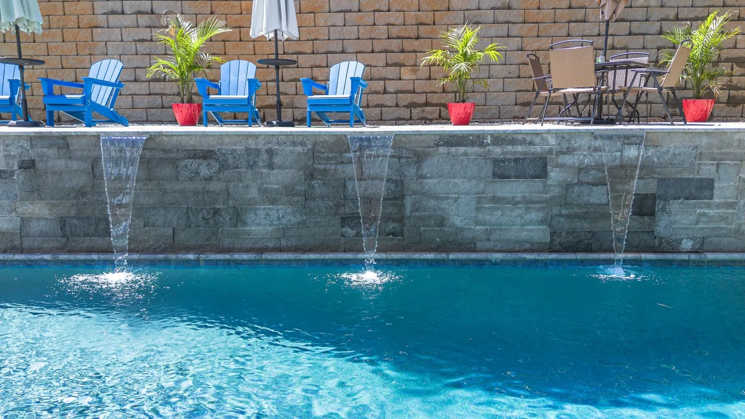 Pool with blue water, stone wall with water features, blue chairs, and small tables.
