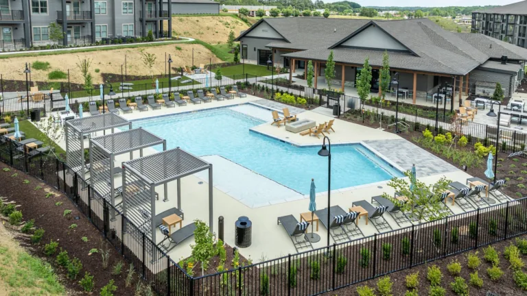 Aerial view of a modern apartment pool with lounge chairs, fenced deck, and clubhouse building