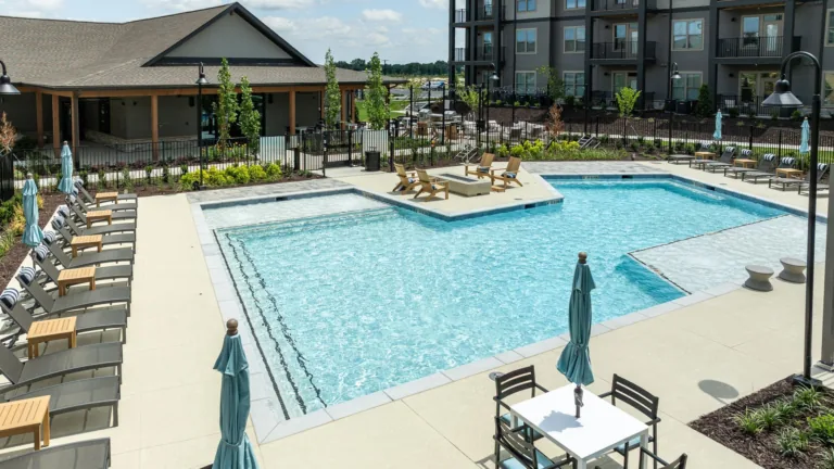 Outdoor apartment pool with lounge chairs, umbrellas, and a hot tub on a sunny day