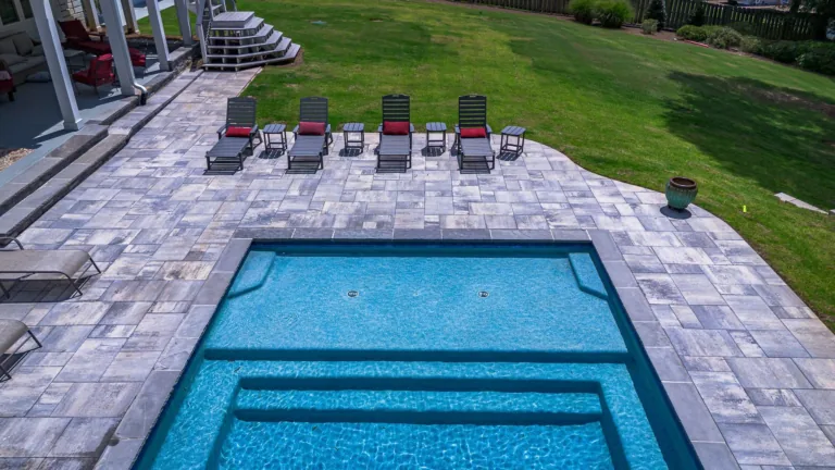 Swimming pool surrounded by gray pavers, with lounge chairs and a green lawn.