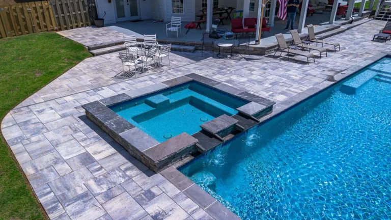 Pool and hot tub with paved patio, white house in background. Blue water, gray pavers, green grass.