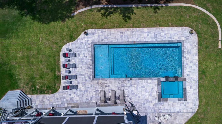Overhead view of a rectangular pool, surrounded by stone patio and grassy yard. Lounge chairs, small hot tub.