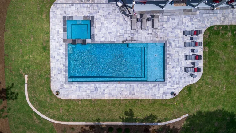 Overhead view of a swimming pool, hot tub, and patio with lounge chairs, surrounded by green grass.