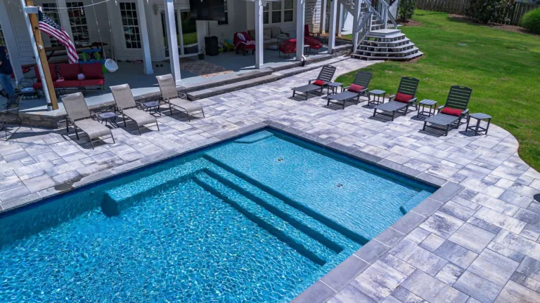 Pool with blue water, steps, and surrounding patio with lounge chairs. Exterior shot with a house in background.