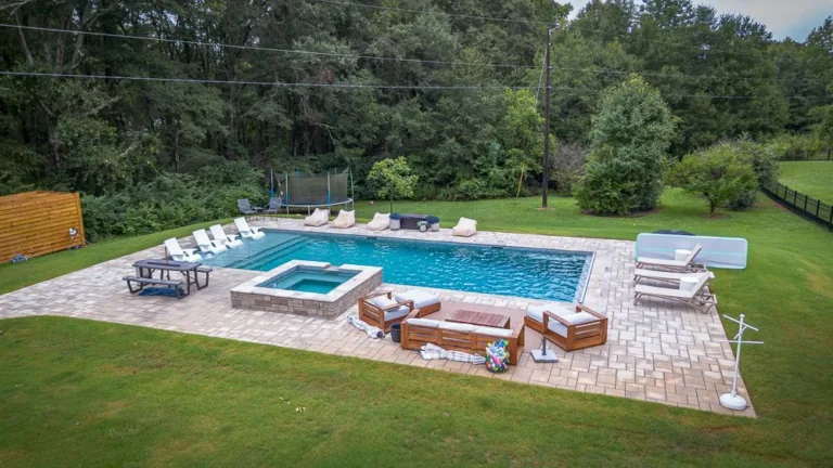 Backyard pool with hot tub, lounge chairs, and seating area surrounded by green grass and trees.