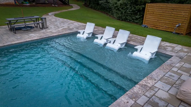 Swimming pool with white lounge chairs in the water. Steps lead into the pool, and a stone patio surrounds it.