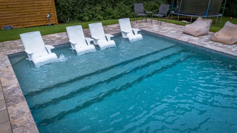 A swimming pool with four white chairs in the shallow end, surrounded by patio and lawn.