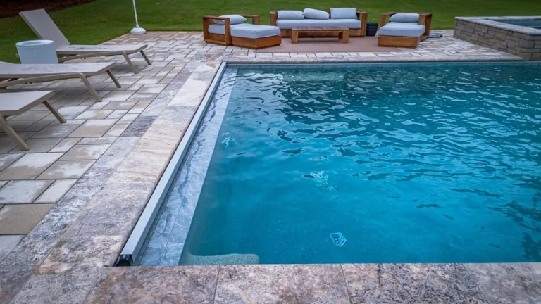 Pool with patio furniture. Light blue water, stone patio, wooden furniture, and green grass.