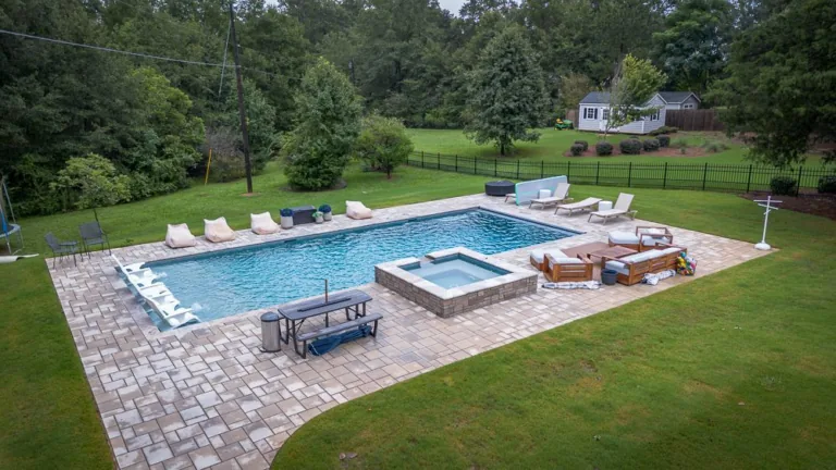 Backyard with pool, hot tub, and lounge area surrounded by green grass and trees.