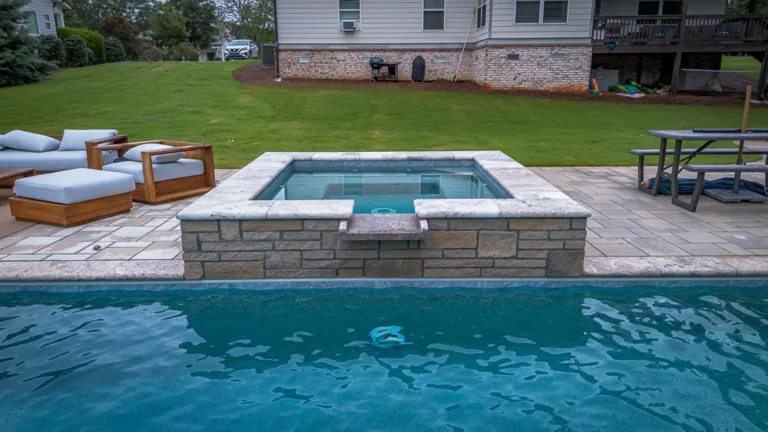 A square stone hot tub next to a pool with seating area and grassy yard in background.