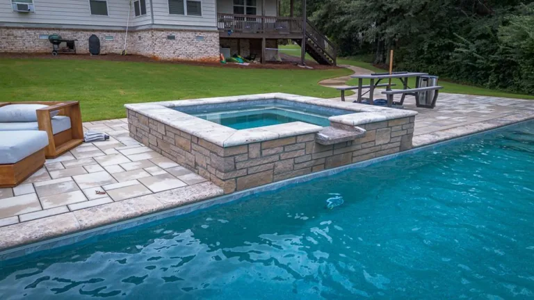 Hot tub next to a pool, on a patio with lawn and house in the background.