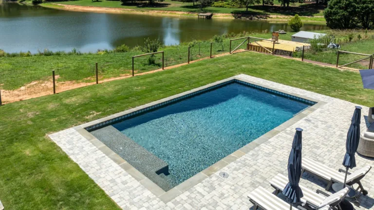 Rectangular blue swimming pool beside a grassy backyard and lake, with patio chairs and umbrellas nearby