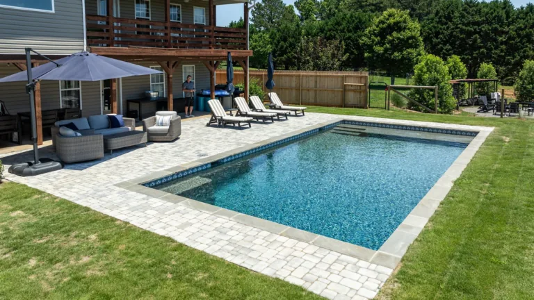 Backyard pool with stone deck, lounge chairs, umbrella, and patio seating beside a house