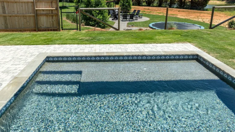 Backyard swimming pool with clear blue water, steps, and stone coping, overlooking a grassy yard.