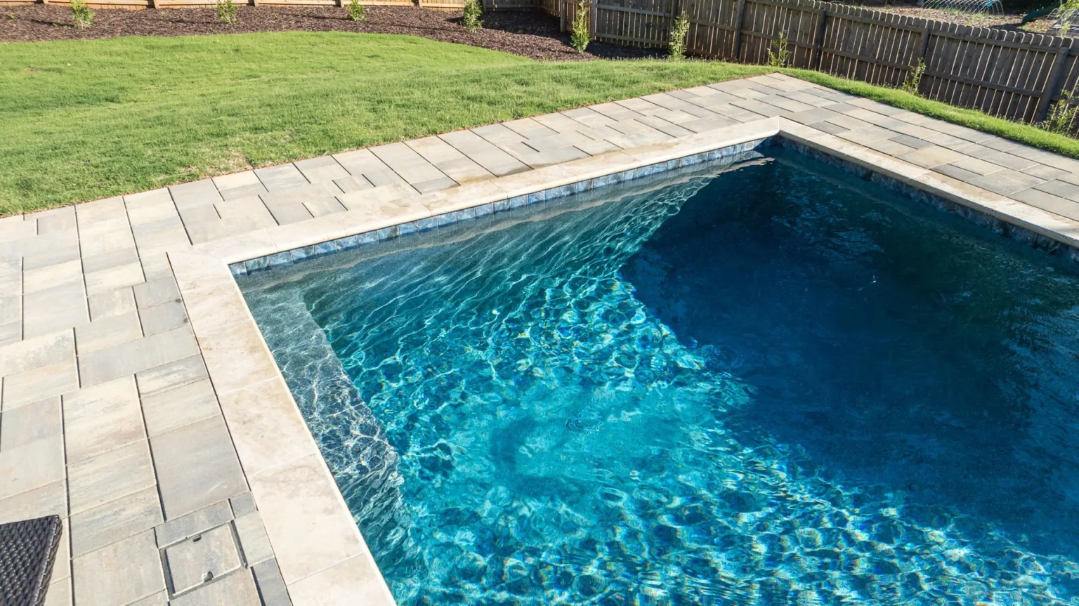 Rectangular backyard swimming pool with blue water and light stone patio surround