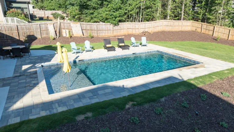 Backyard swimming pool with lounge chairs, stone deck, and lawn in bright sunlight