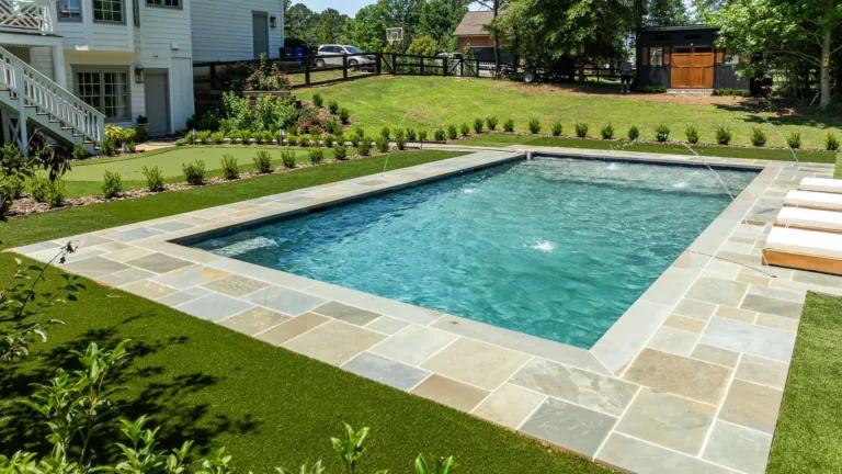 Rectangular backyard swimming pool with stone coping, surrounded by green lawn and a house in the background