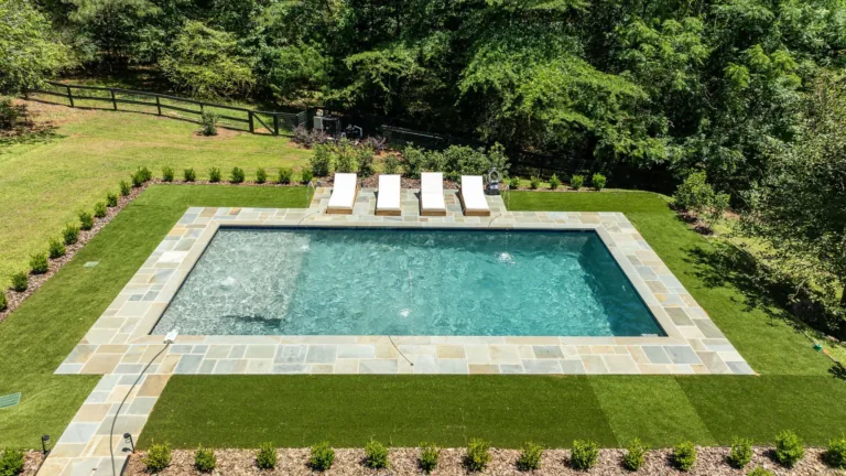 Rectangular backyard swimming pool bordered by stone, with four lounge chairs and green lawn and trees around it