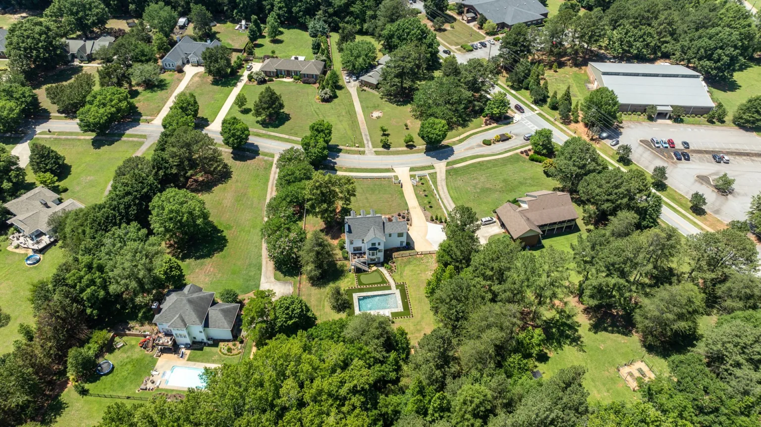 Aerial view of a tree-lined neighborhood with houses, roads, and a swimming pool on a sunny day