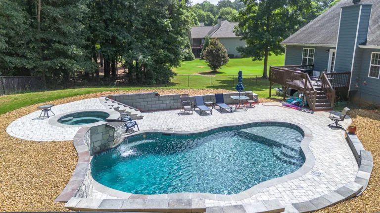 Backyard pool with a spa, waterfall, and stone patio, near a house and trees.