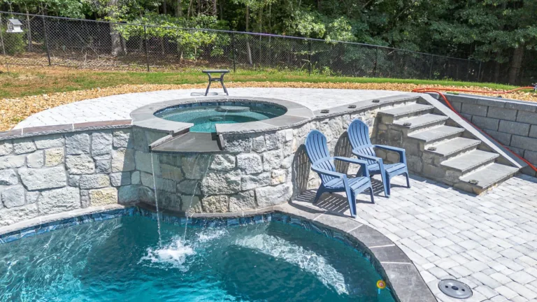 Stone pool with a waterfall feature, hot tub, blue chairs, and paved patio steps.