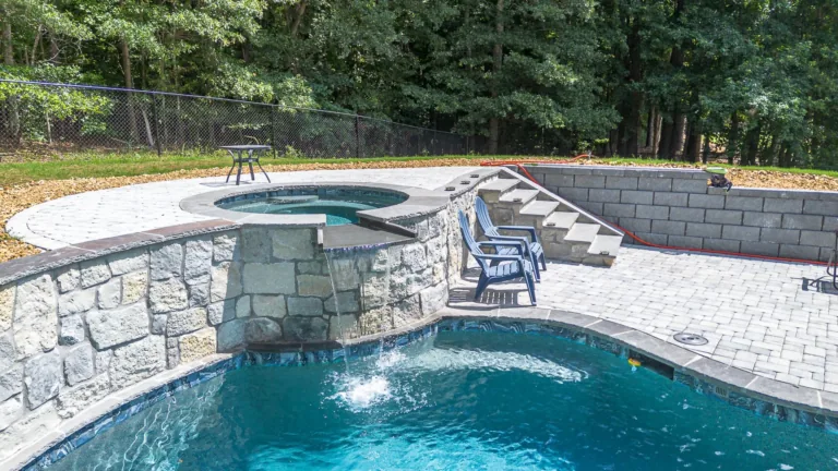 Pool with a spa, stone walls, steps, and lounge chairs in a backyard setting.