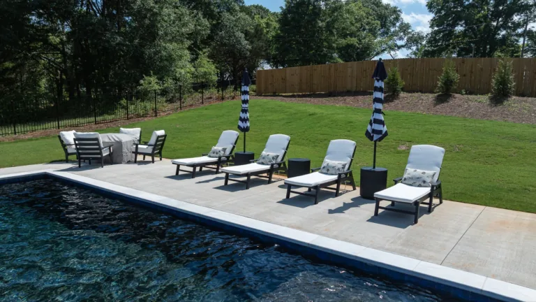 Backyard pool deck with lounge chairs, umbrellas, and a fenced lawn beyond the water