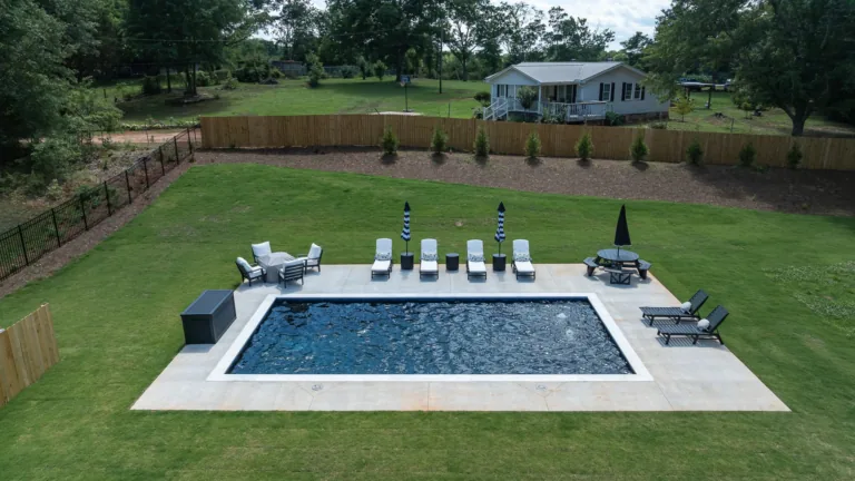 Backyard with a rectangular in-ground pool, patio chairs, and a covered seating area on a green lawn.