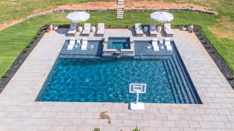 Outdoor pool with loungers, umbrellas, and a basketball hoop, surrounded by stone patio and lawn.