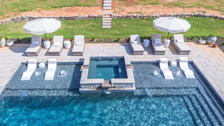 Luxury pool with loungers, umbrellas, and a spa area. Blue water, green grass, sunny day.