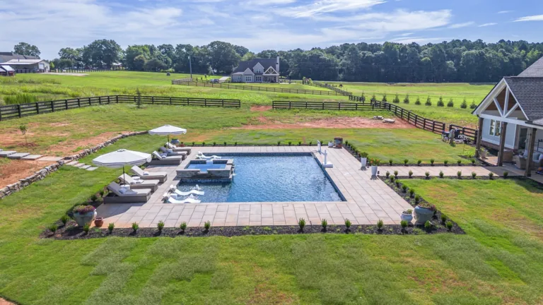 Square swimming pool surrounded by green grass, lounge chairs, and a small building; set in a large yard near trees.