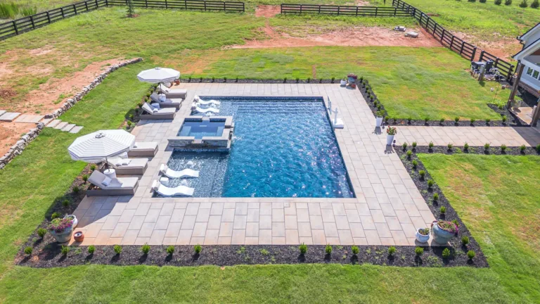 Aerial view of a rectangular swimming pool with a hot tub. White lounge chairs and umbrellas are poolside on a grassy lawn.