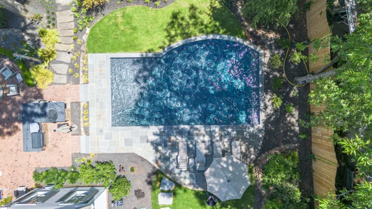 Overhead view of a backyard with a pool, patio, lawn, fence, and trees on a sunny day.