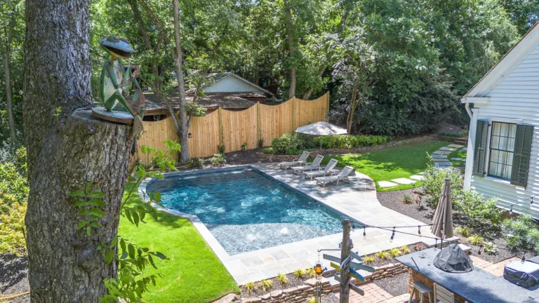 Backyard pool with lounge chairs, small fence, and house. Lush greenery and tree in the foreground.