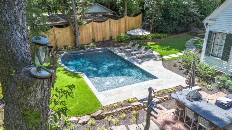 Backyard with pool, lounge chairs, and a white house. Green grass and a bamboo fence surround the pool.