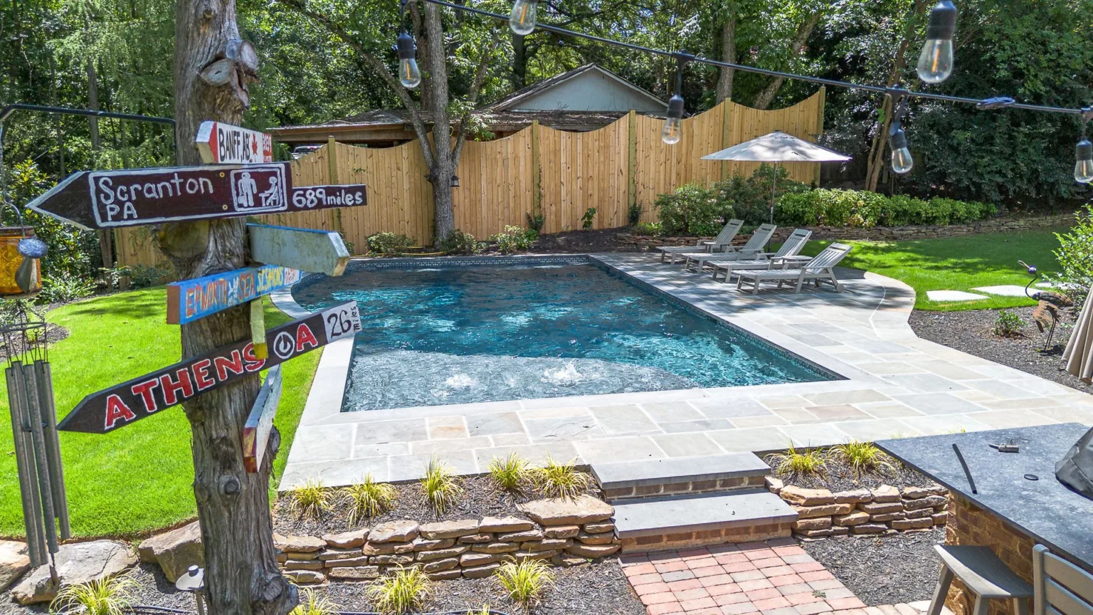 Backyard with pool, wooden fence, string lights, signs, and lounge chairs.