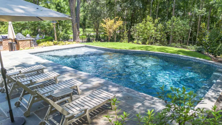 A backyard pool with white lounge chairs. Trees surround the pool with a stone patio and umbrella.