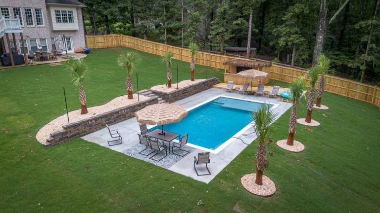 A backyard pool with patio furniture, landscaping, and palm trees, bordered by a wooden fence.