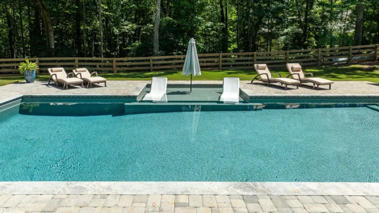Outdoor swimming pool with lounge chairs, white umbrellas, and a wooden fence amid trees